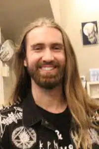 A man with long blonde hair and a beard, wearing a black shirt with a logo, smiling confidently against a blurred background.