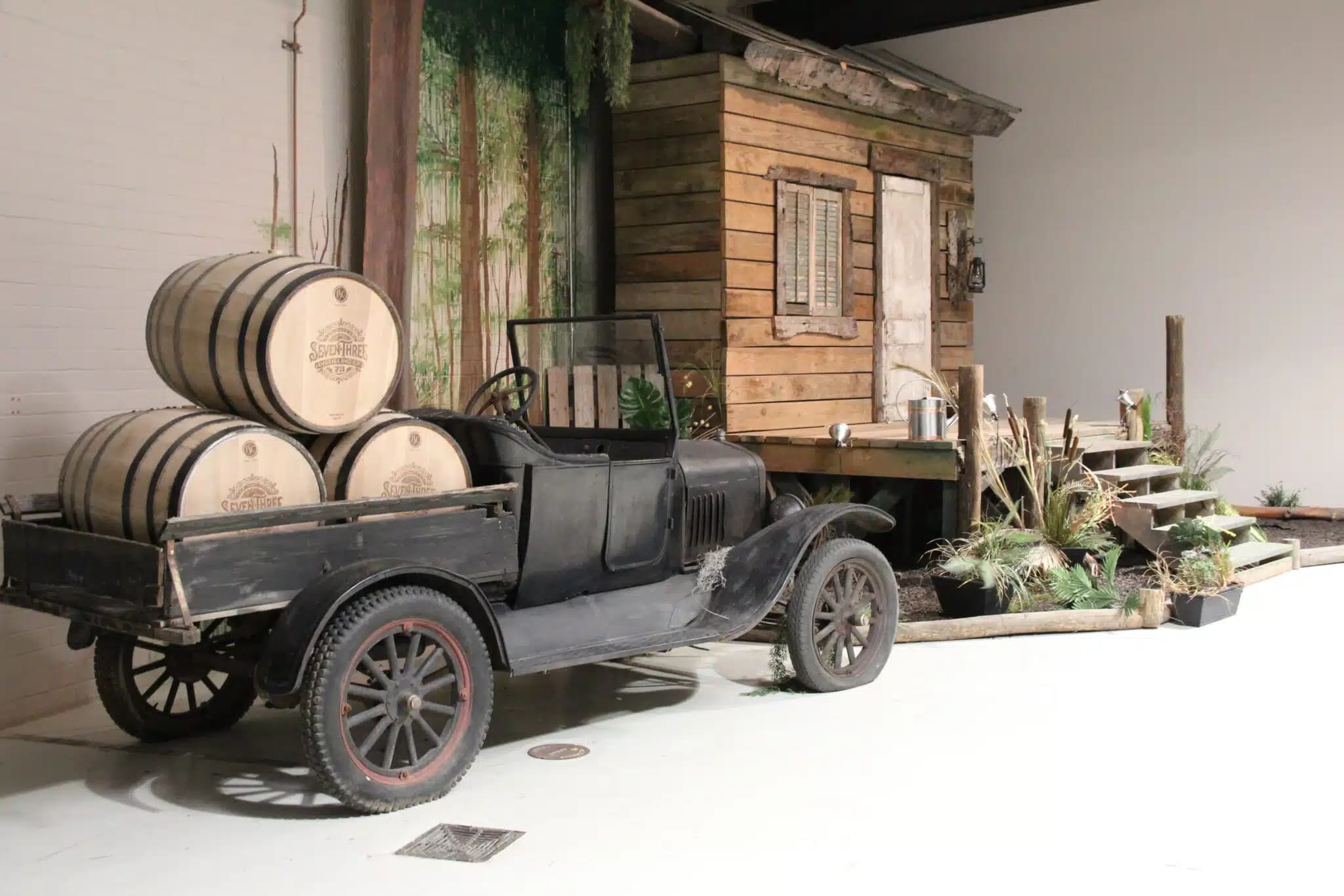 A vintage black truck loaded with wooden barrels parked beside a rustic wooden cabin with a small porch and stairs, surrounded by plants in an indoor exhibit