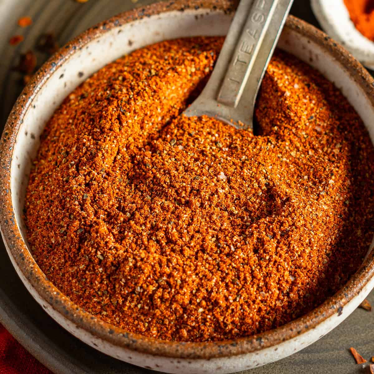 A bowl of orange colored Cajun seasoning with a metal spoon inside.